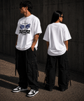 Two people wearing white t-shirts with graphic designs, standing against a dark wall.
