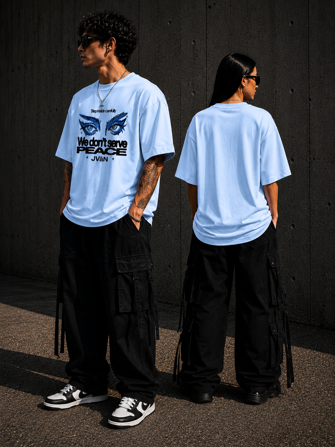 Two people wearing light blue t-shirts with a graphic design and text, standing against a dark wall.