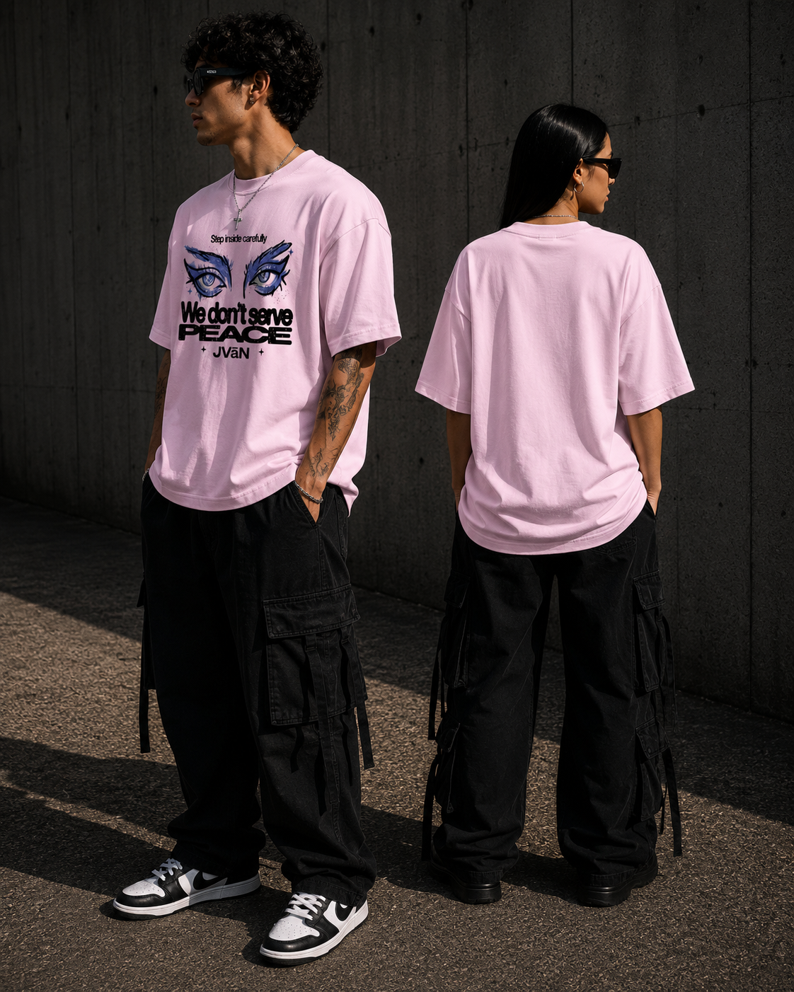 Two people wearing pink t-shirts with a graphic design and text, standing against a dark concrete wall.