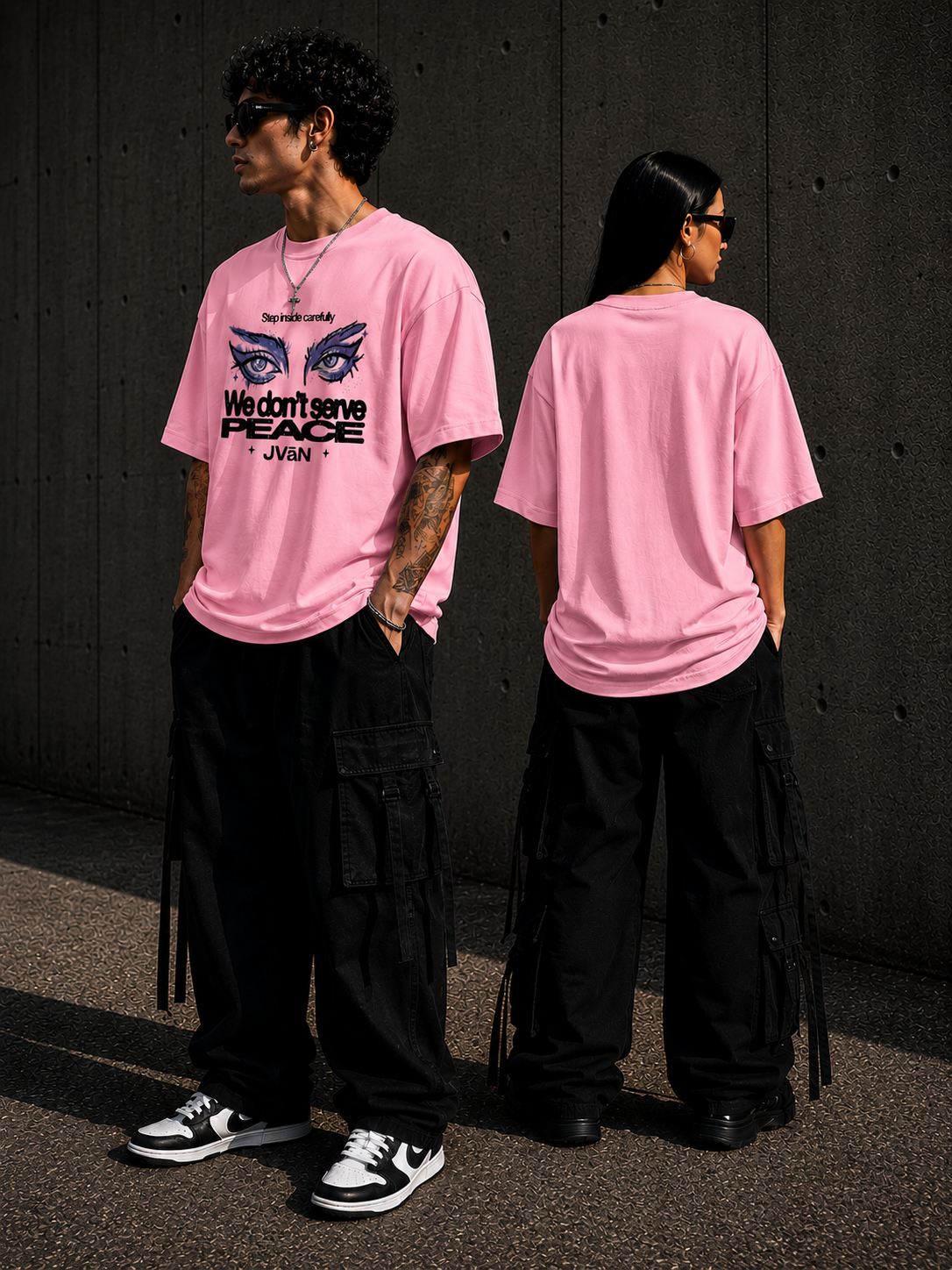 Two people wearing pink t-shirts with a graphic design and text, standing against a dark wall.