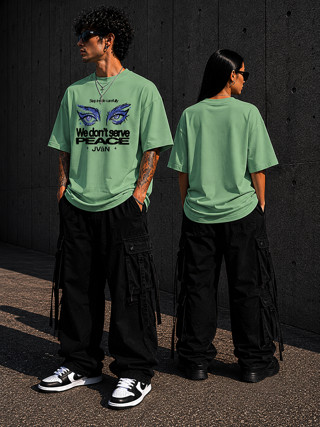 Two people wearing green t-shirts with a graphic design and text, standing against a dark wall.