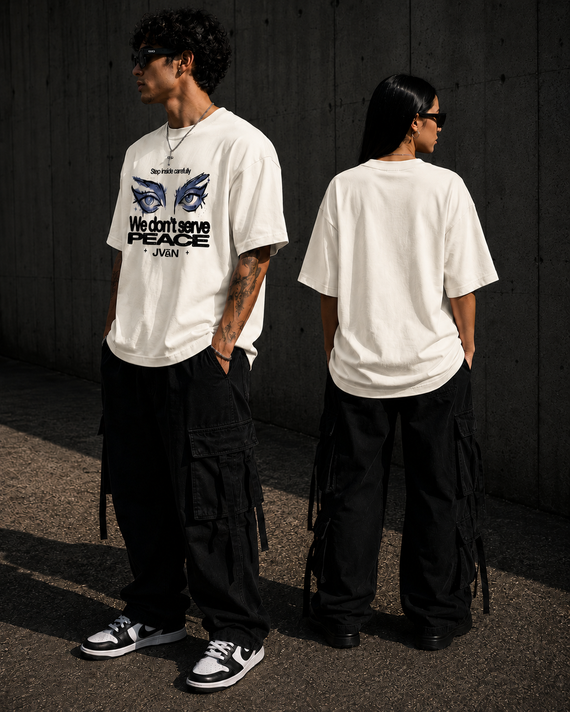 Two people wearing white t-shirts with graphic designs, standing against a dark concrete wall.